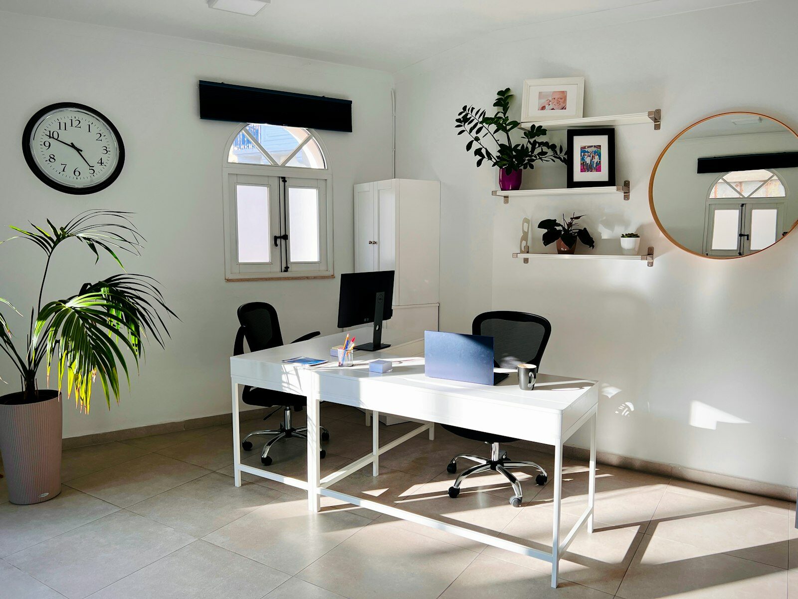 A bright office with two white desks, an open laptop, a monitor and a large potted palm, lit by sunlight through an arched window.