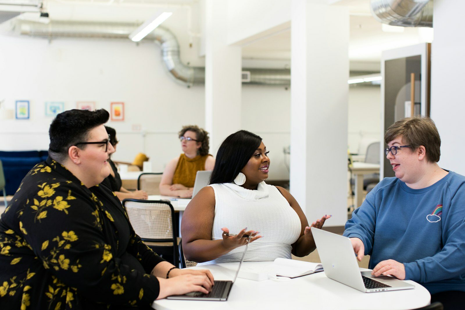 Three colleagues talking around a white table with laptops open in a bright open-plan office.