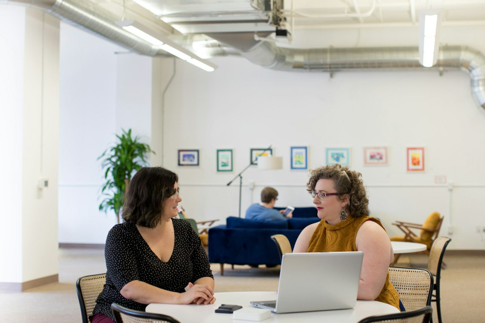 Two colleagues in conversation at a small round table, one with a laptop, in a bright open-plan office.