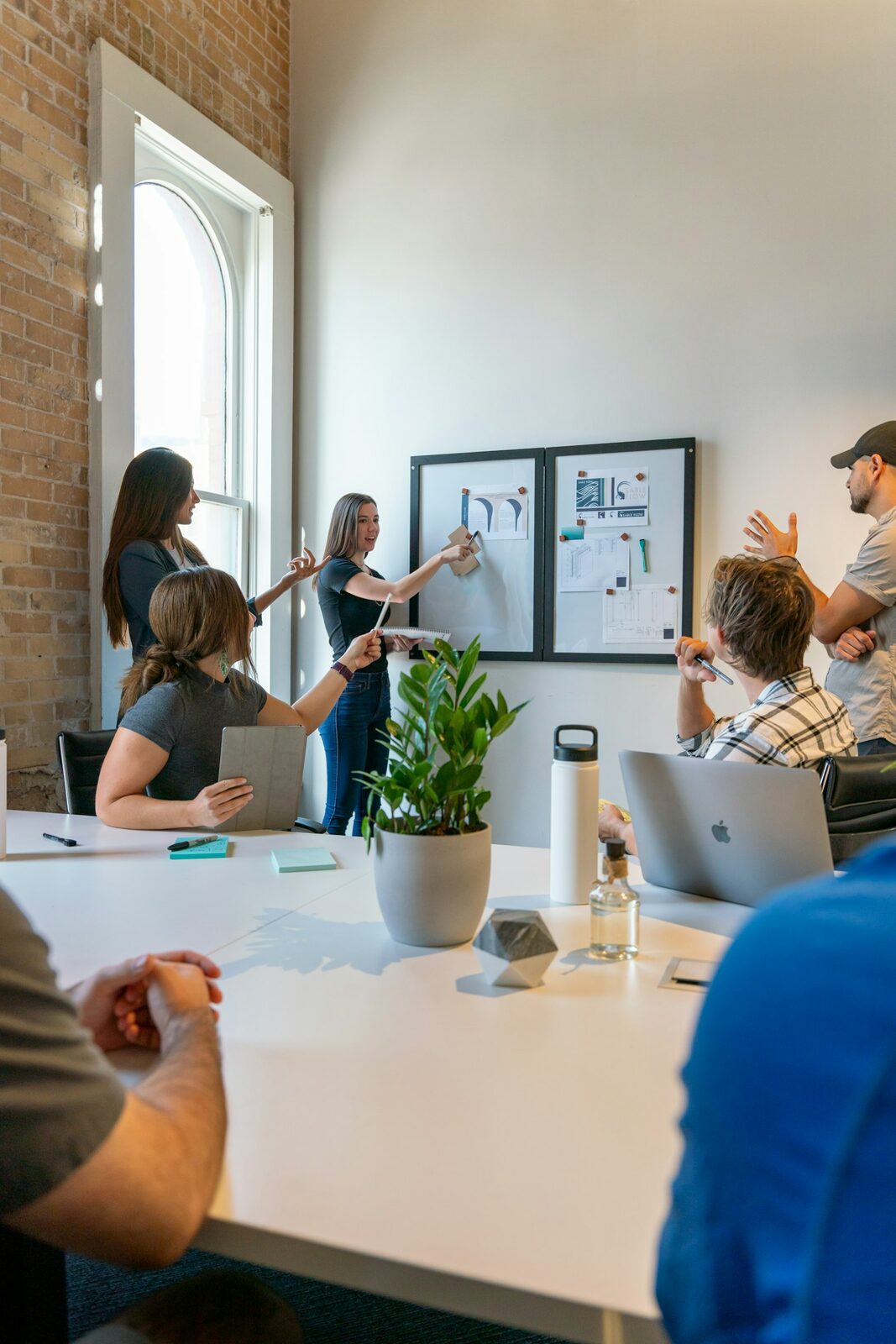 A group of colleagues gathered around a whiteboard in a meeting room, one standing and pointing at a pinned diagram.